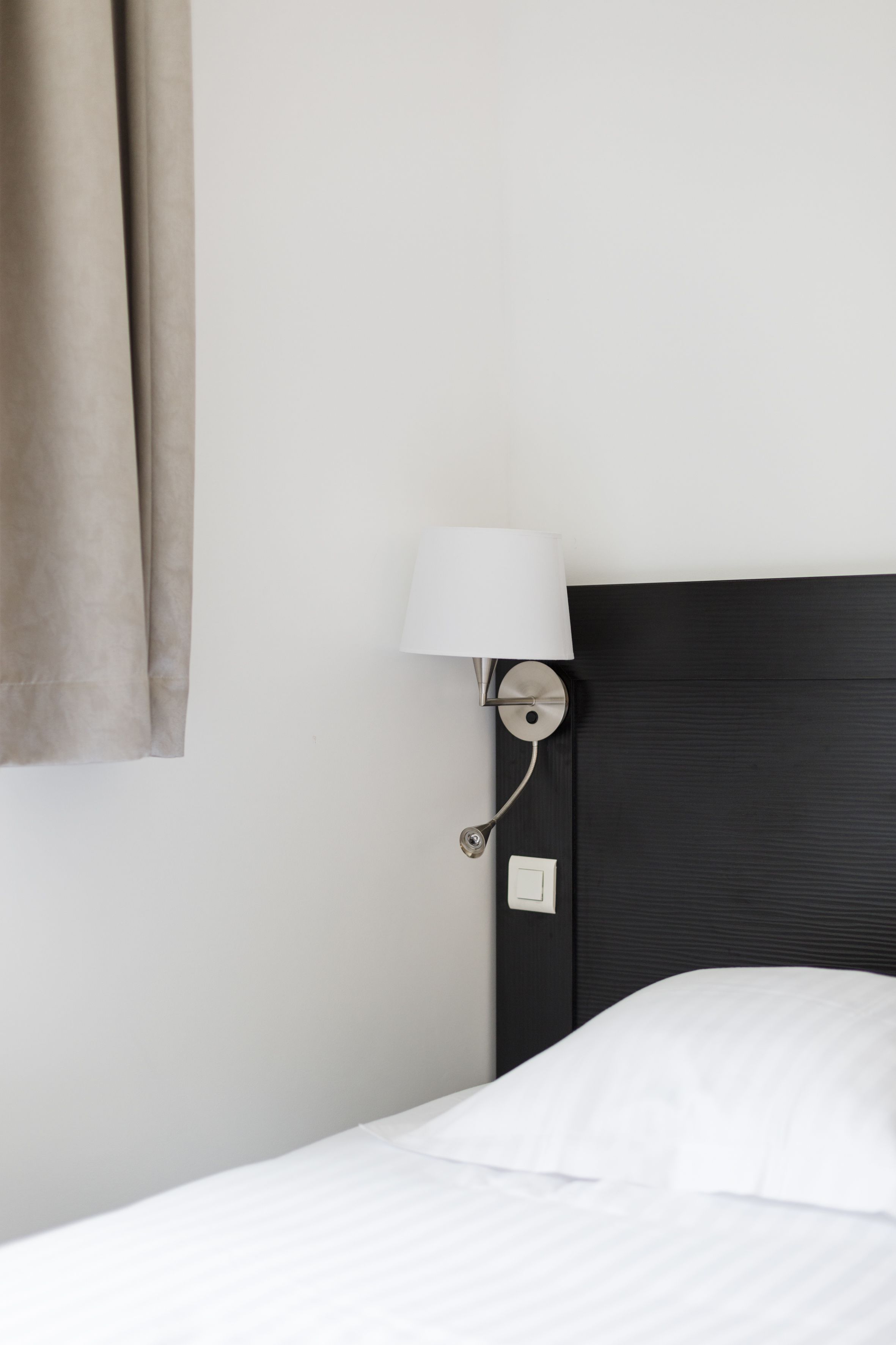 A hotel room with a bed, a wall-mounted lamp, and a curtain.