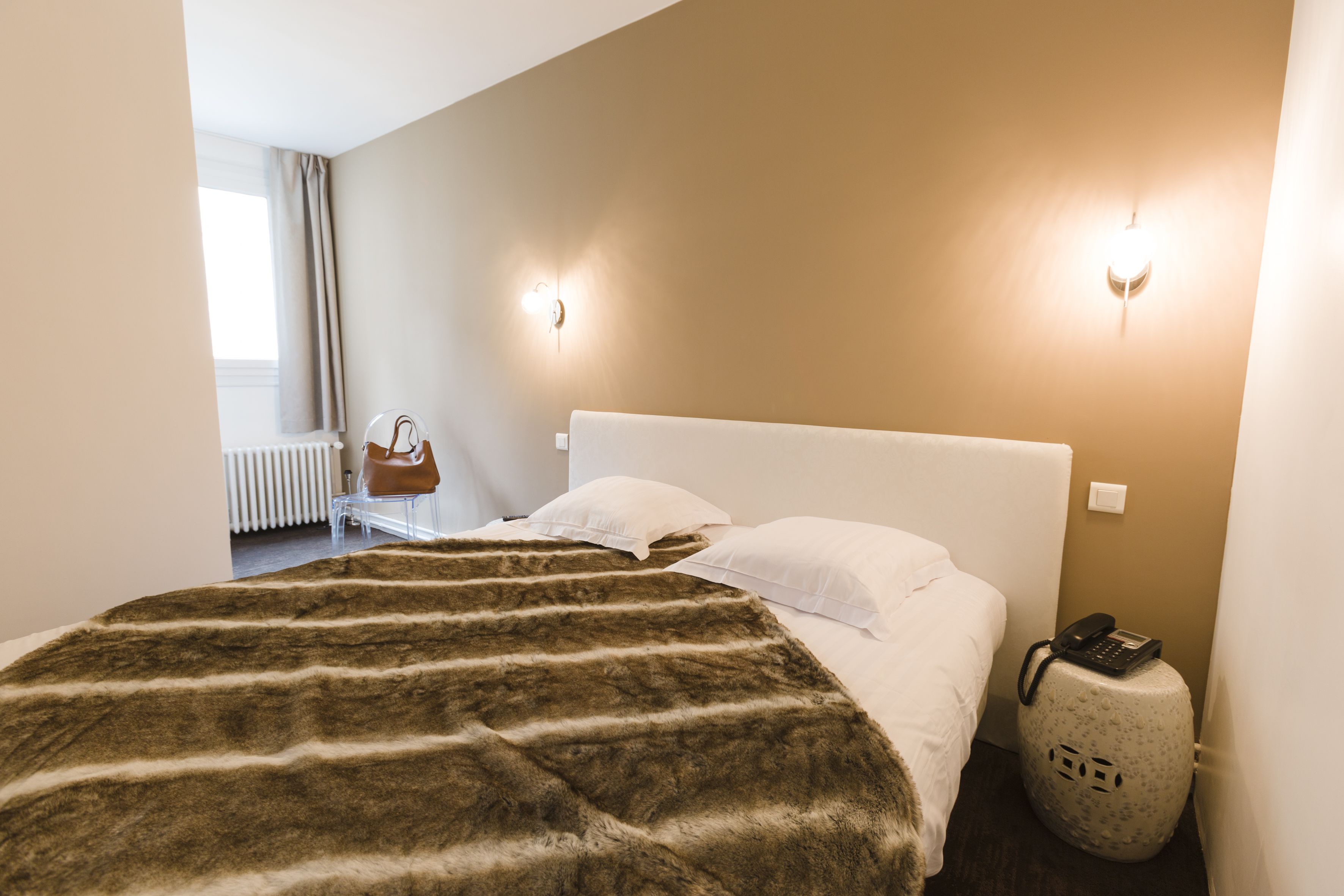 A hotel room with a large bed, a white headboard, a fur throw, a handbag, a suitcase, and a wall-mounted phone.