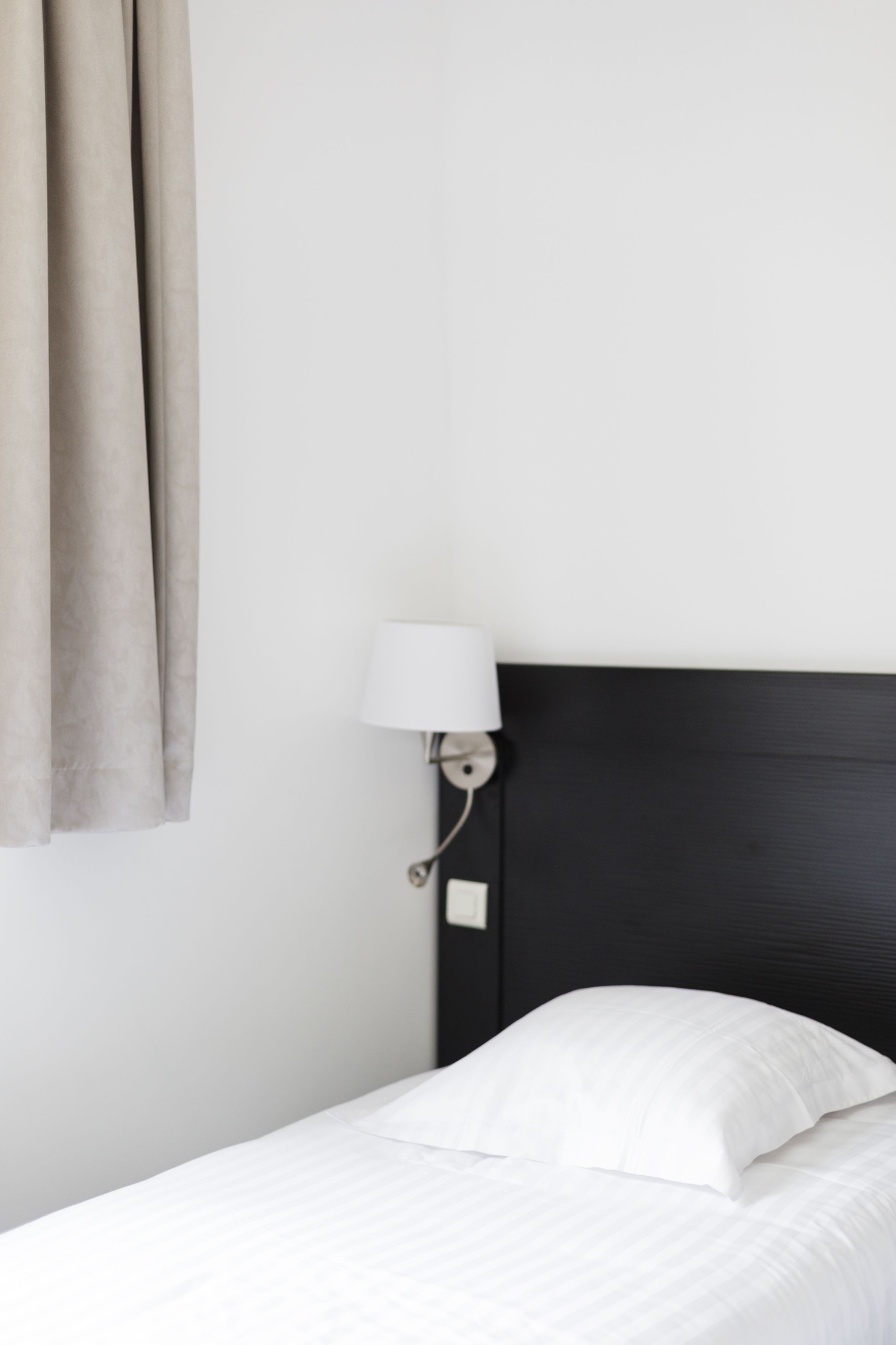 Una cama con ropa de cama blanca, cabecera oscura y lámpara fijada a la pared.