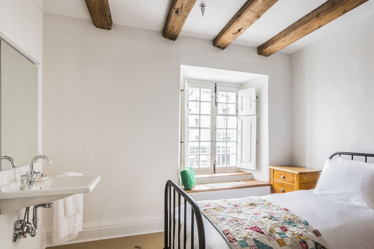 A bedroom with a bed, a window, a wooden ceiling, a bedside table, and a bathroom with a sink and mirror.