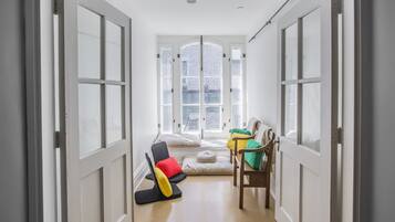 A narrow hallway with a large window at the end, leading to a room with a bed and two chairs.