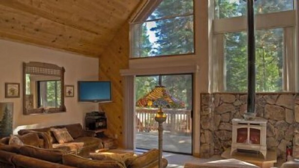 A cozy cabin interior with a stone fireplace, wooden ceiling, and large windows offering a view of the outdoors. - St. Croix (Truckee)