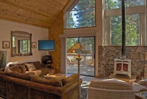 Interior acogedor de una cabaña con chimenea de piedra, techo de madera y ventanas amplias que dan al exterior.