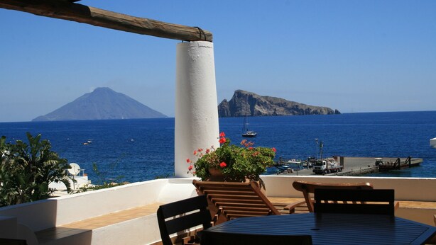 A terrace with a table and chairs, overlooking a body of water with boats and a mountain in the background. - Hotel Tesoriero (Panarea)