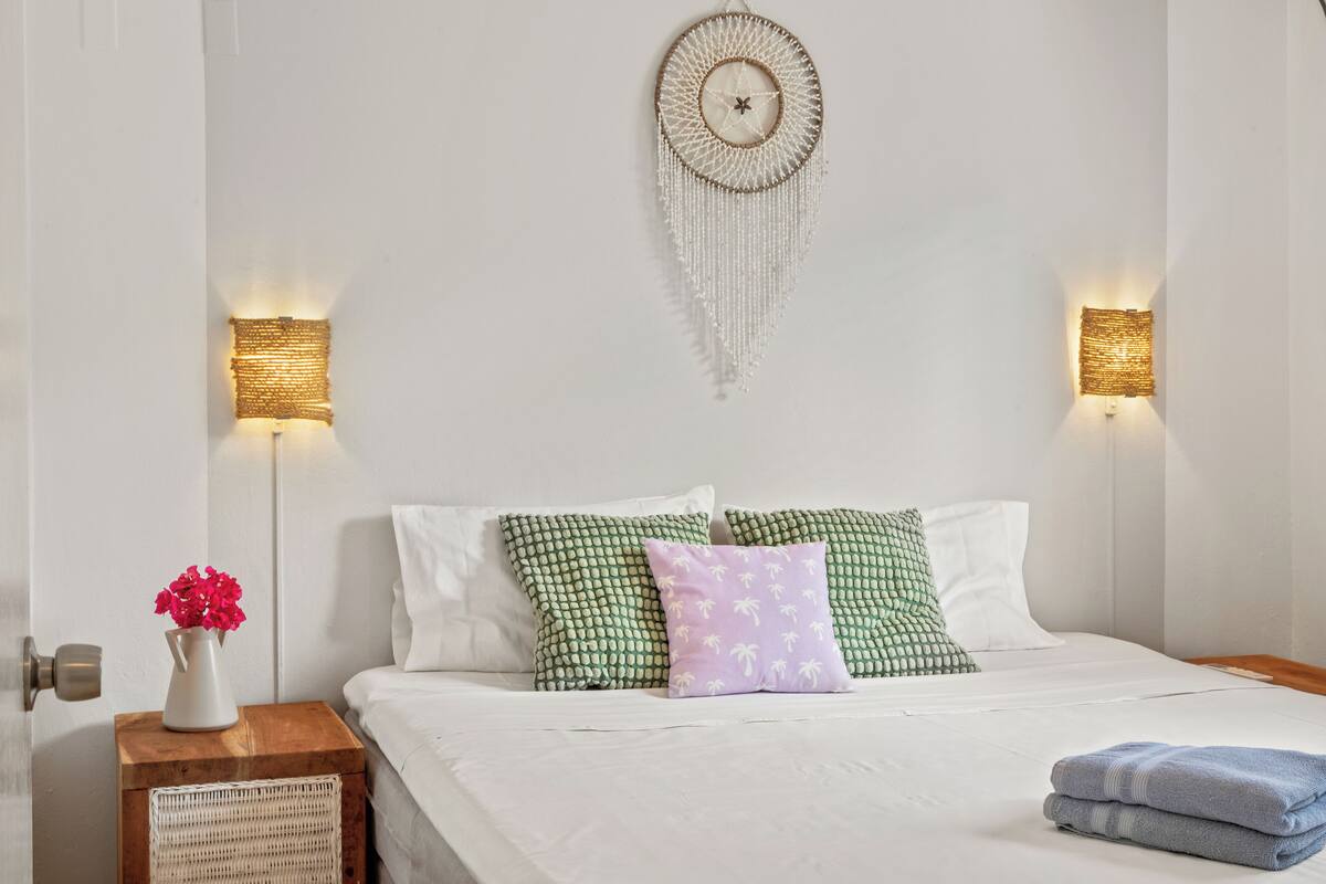 A neatly made bed with white bedding and decorative pillows, a wooden nightstand with a vase of flowers, and wall-mounted light fixtures.