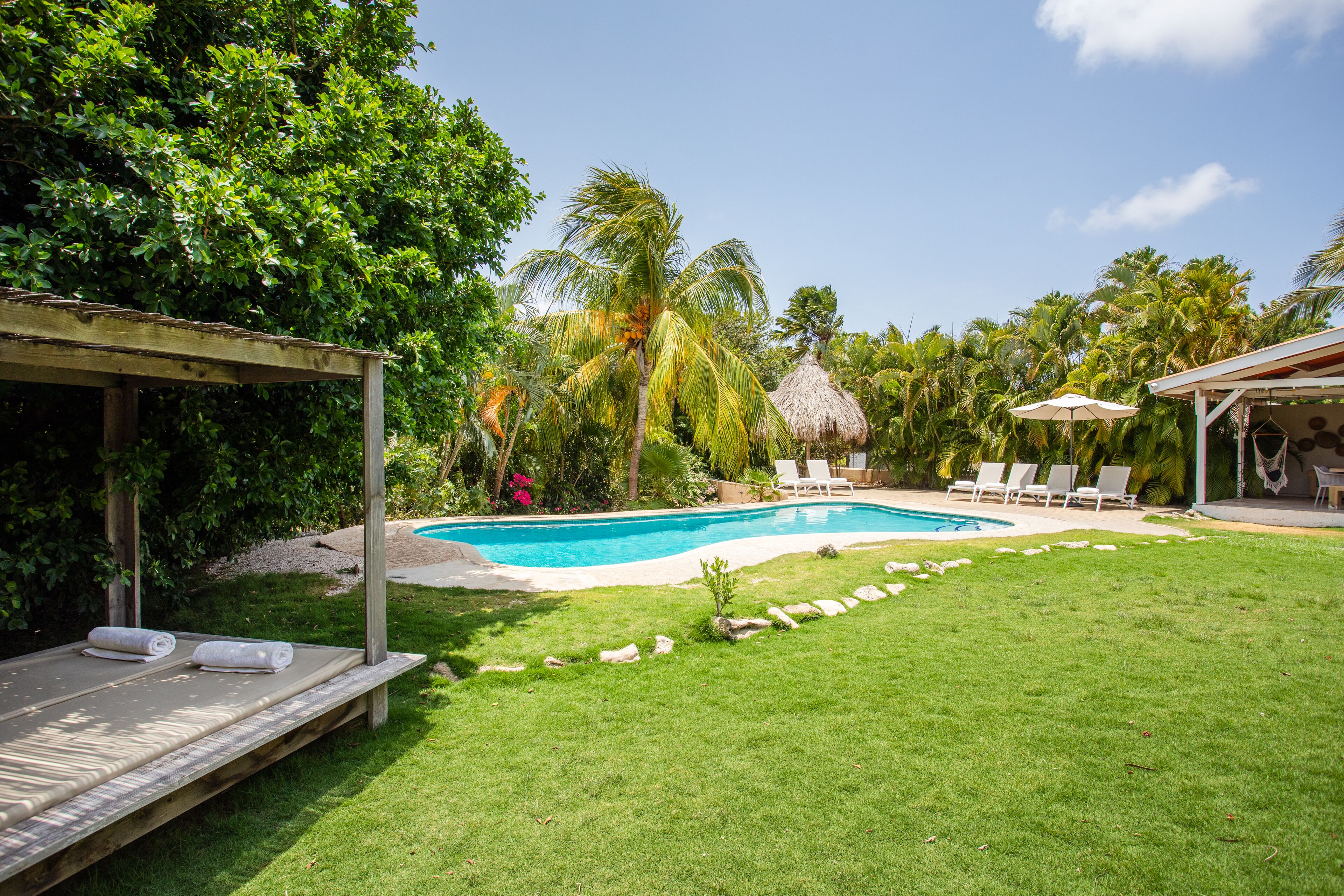 A swimming pool surrounded by a grassy lawn, with lounge chairs and a thatched-roof structure.