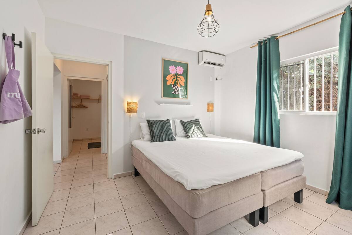 A bedroom with a bed, a hanging light fixture, a wall-mounted air conditioner, and a window with curtains.