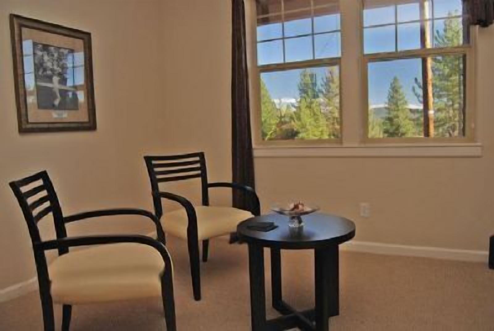 A room with two chairs and a small table, a framed picture on the wall, and a window with a view of trees.
