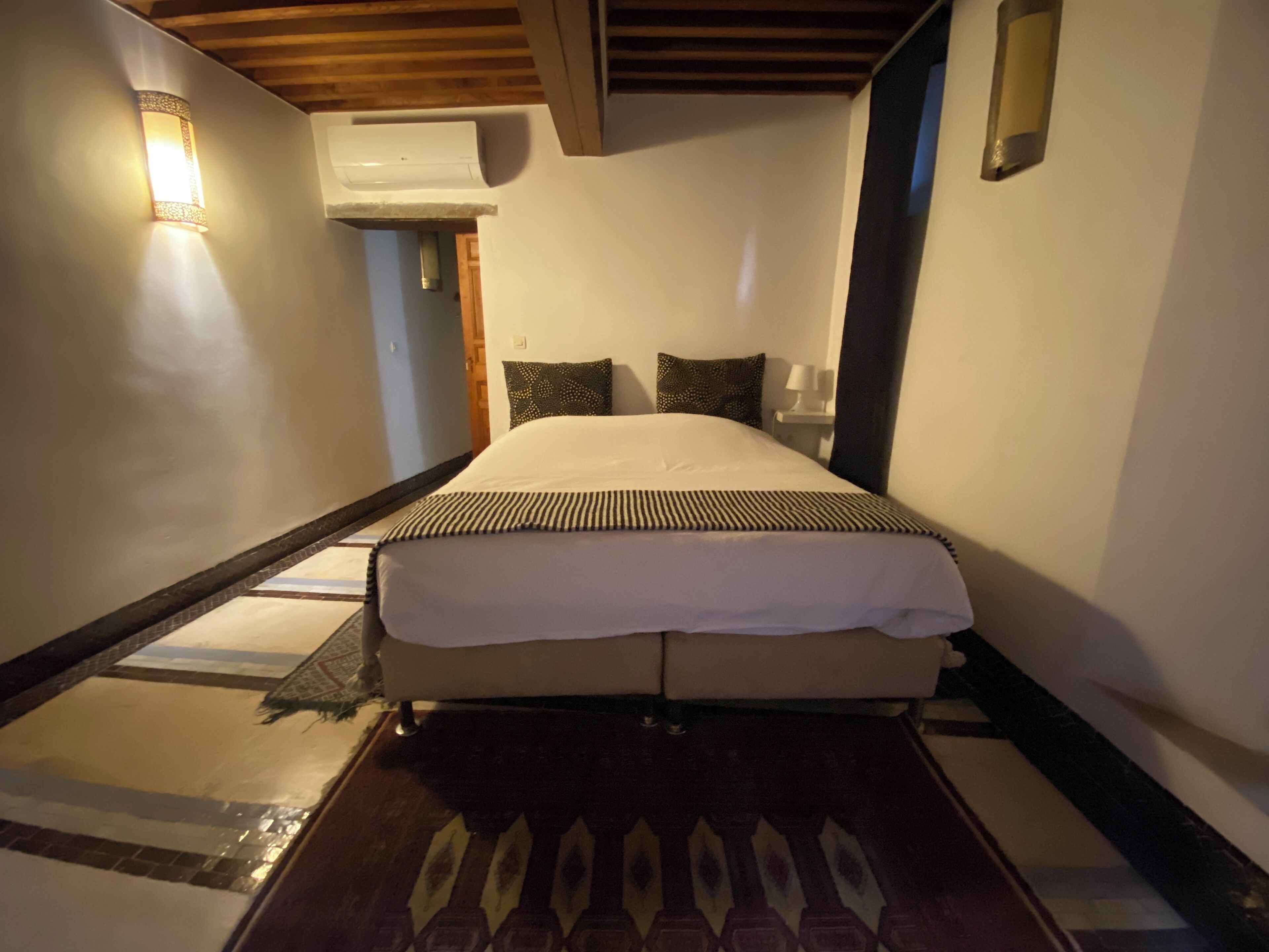 Habitación de hotel con cama, aire acondicionado de pared, mesita de noche con lámpara y suelo estampado.