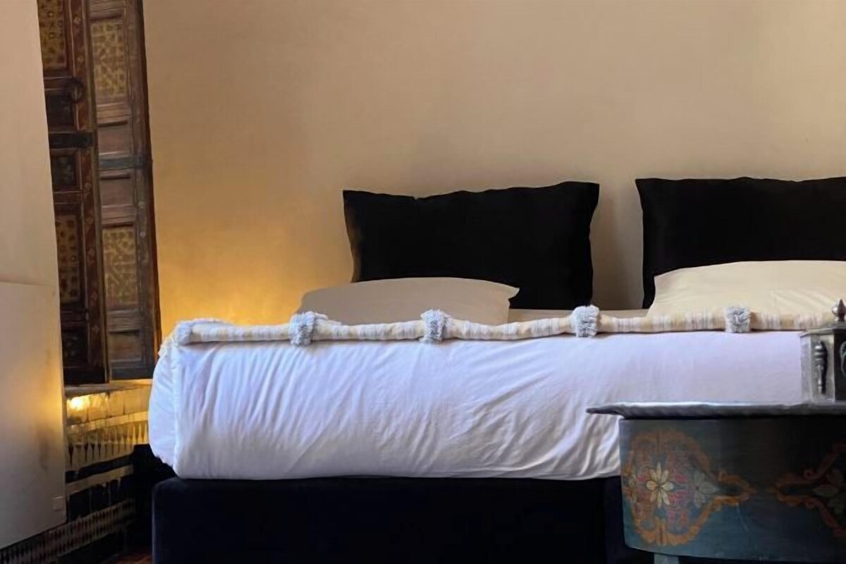 A bed with white bedding and black pillows, a small table with a decorative item, and a patterned wall.