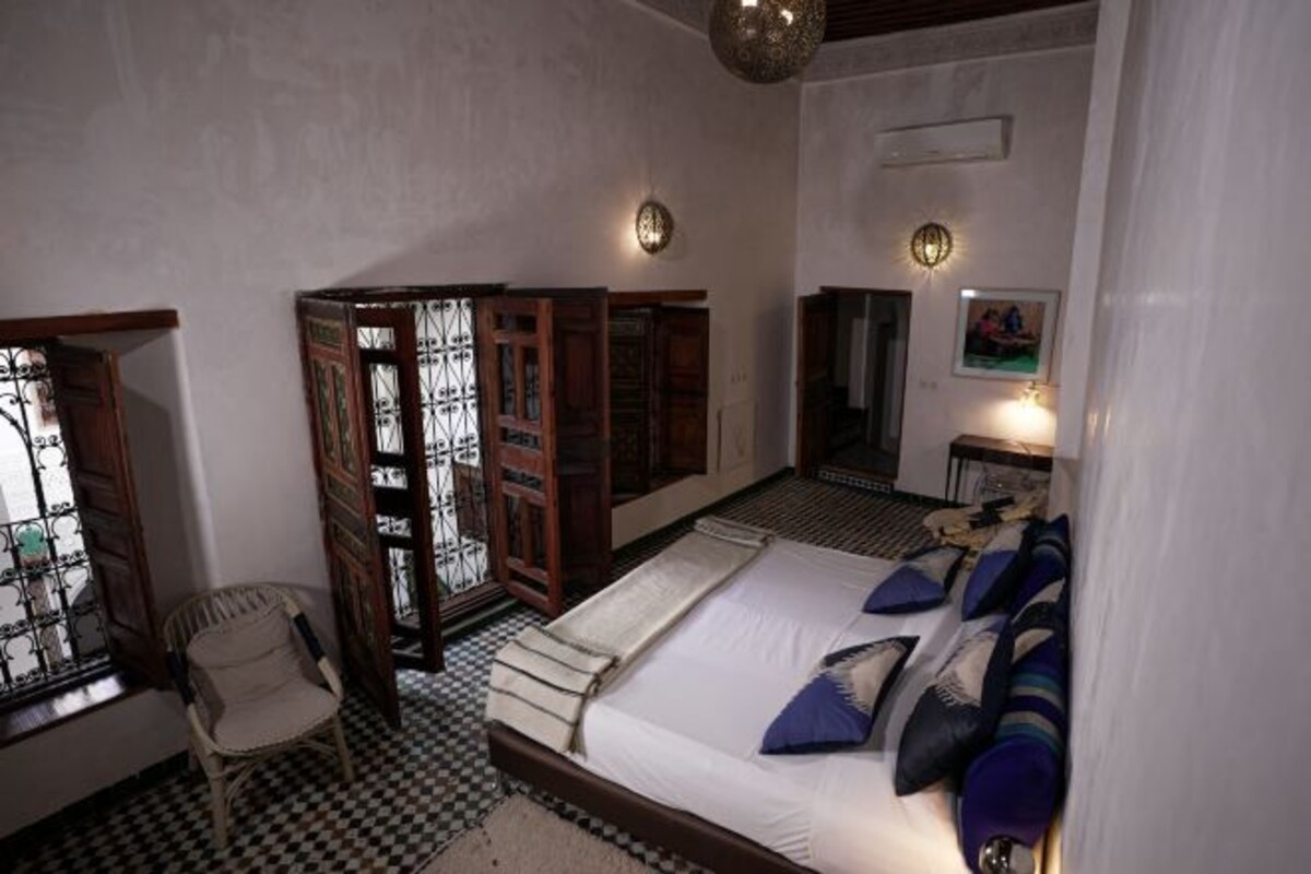 A bedroom with a checkered floor, a bed with blue and white bedding, a wooden chair, and a window with metal bars.