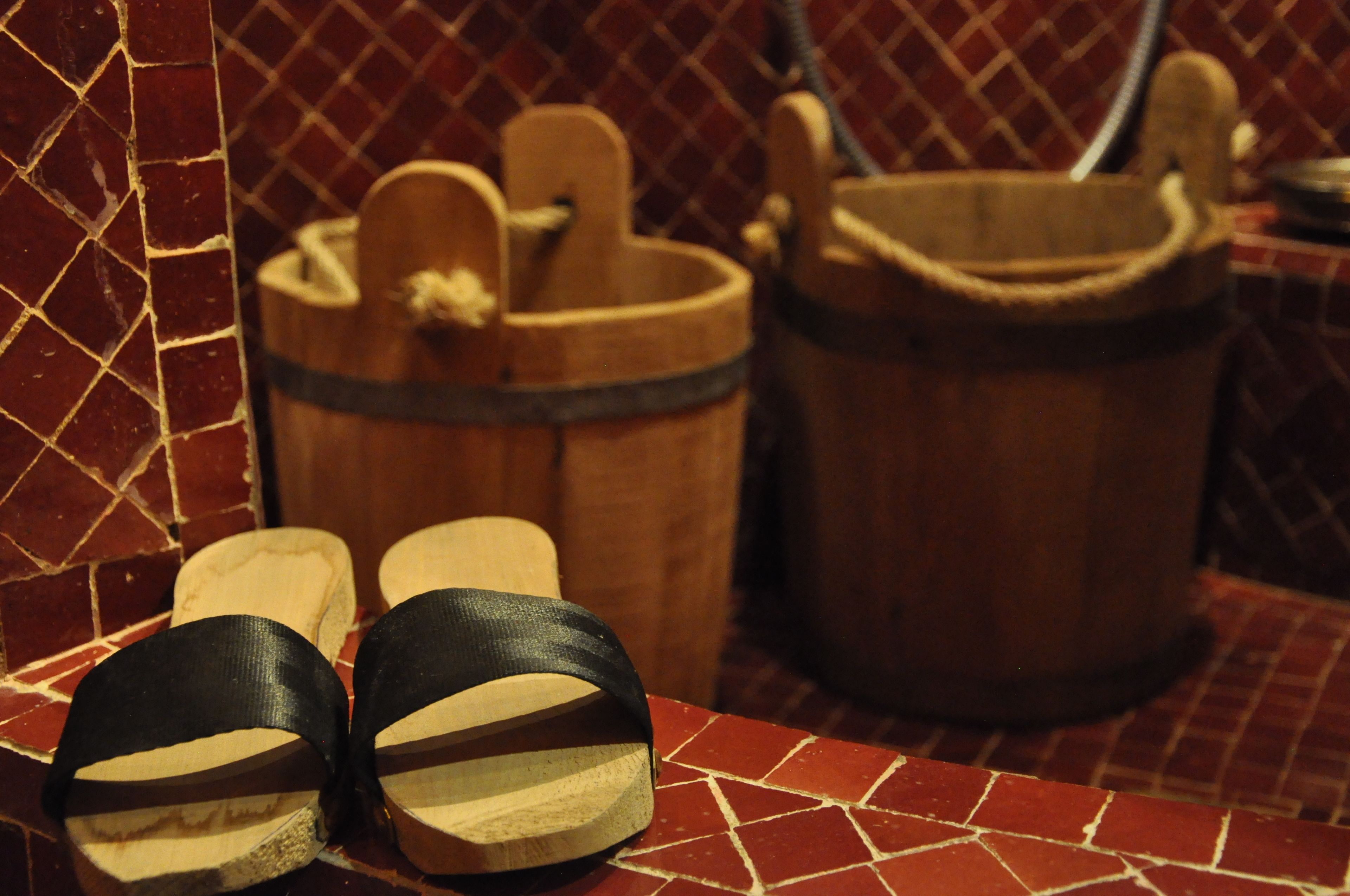 A bathroom with a tiled floor, a wooden bucket, and a pair of black sandals.