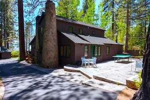 A two-story house with a stone chimney, a patio with outdoor furniture, and a forested backdrop.