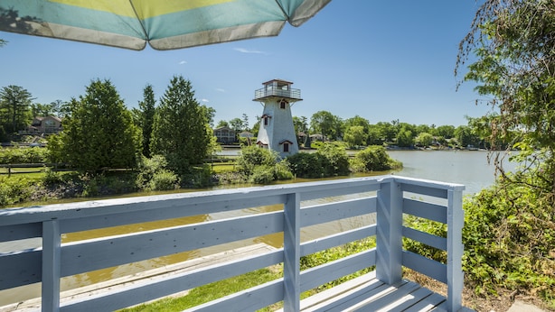 A wooden deck with a white railing overlooking a lake, a lighthouse, and a striped umbrella. - Hollywood Cottages (Wasaga Beach)