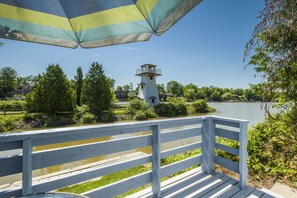 A wooden deck with a white railing overlooking a lake, a lighthouse, and a striped umbrella. - Hollywood Cottages (Wasaga Beach)