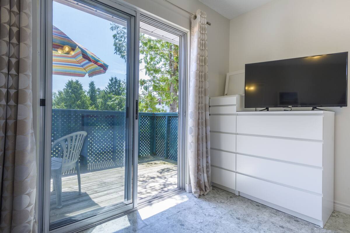 A room with a sliding glass door leading to an outdoor area with a patio and a white chair. A flat-screen TV is mounted on the wall.