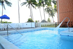 A swimming pool with a water feature, surrounded by palm trees and a covered area with white chairs. - Guaxuma Praia Hotel (Maceió)