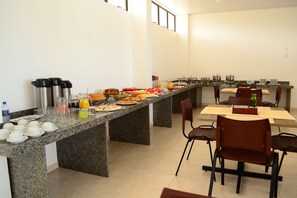 A breakfast buffet with various food items and beverages, a long counter, and several chairs around a table. - Guaxuma Praia Hotel (Maceió)