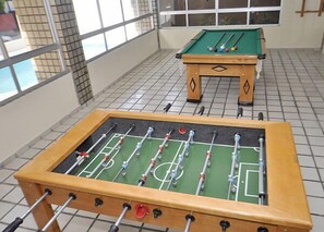 Uma mesa de futebol de mesa e uma mesa de bilhar numa sala com chão em azulejos e janelas amplas.