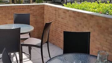 Un patio al aire libre con una mesa de vidrio redonda y cuatro sillas, un muro de ladrillo y una pequeña área ajardinada.