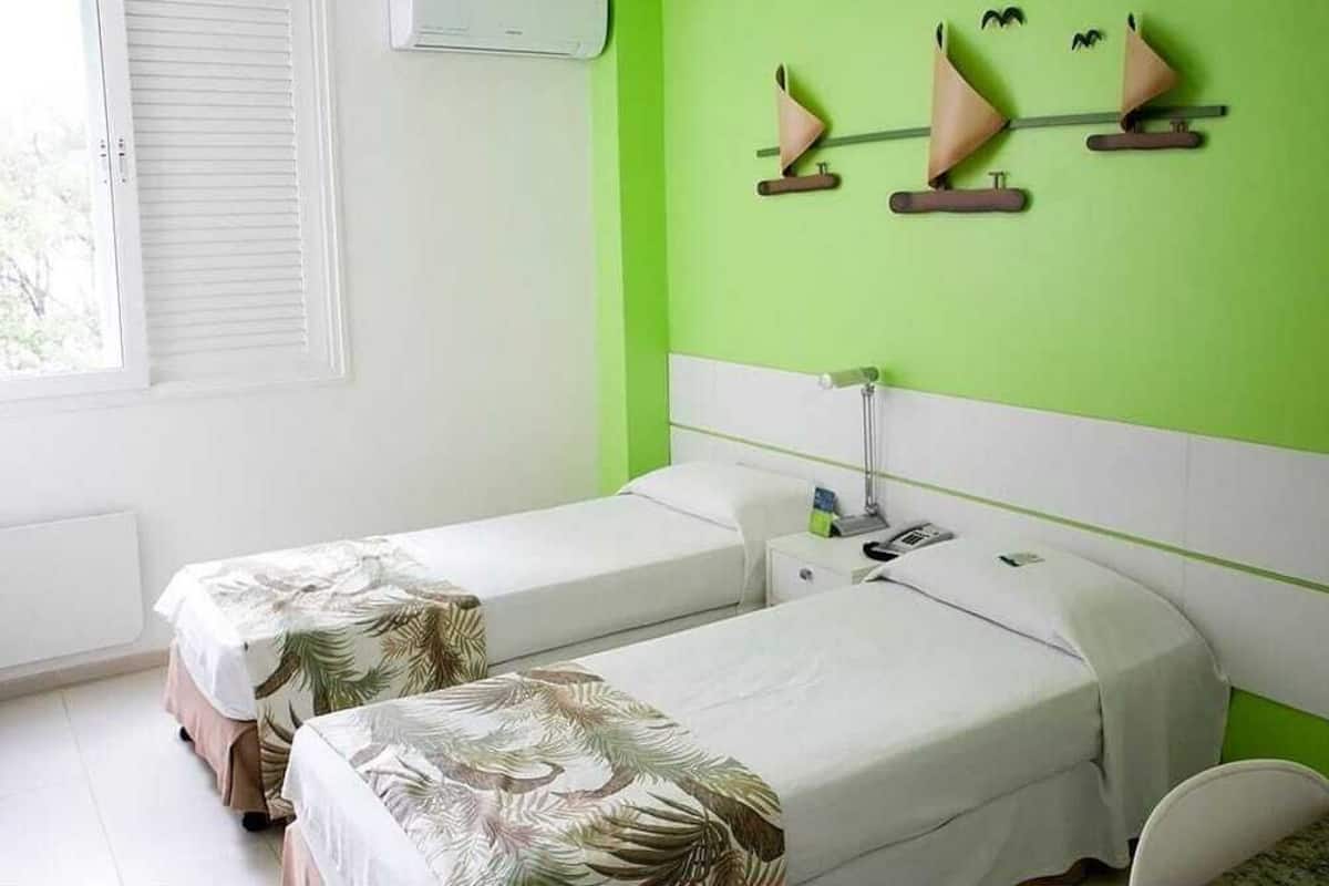 Een kamer met twee bedden, een raam met zonwering, een groene wand en een wandplank met decoratieve objecten.