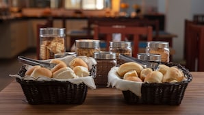 Two wicker baskets filled with bread rolls on a wooden table, with jars of condiments in the background. - Hotel Ciribaí (Maceió)