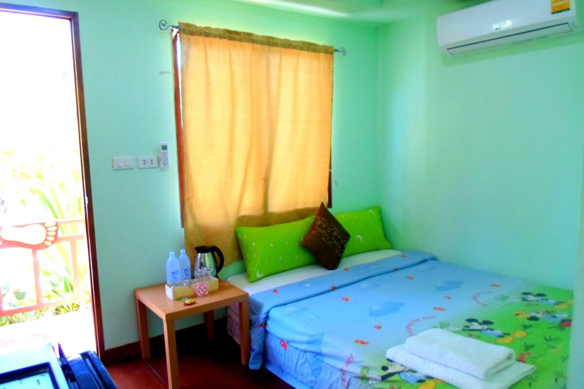 A small, brightly lit room with a bed, a nightstand, a window with curtains, and an air conditioning unit.