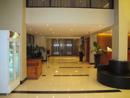 Un vestíbulo de hotel con mostrador de recepción, refrigerador, plantas en macetas y una escalera que lleva a un nivel superior.