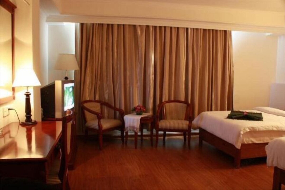 A hotel room with a wooden floor, a bed, two chairs, a desk with a lamp, a television, and a curtain.