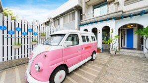Ein pinkfarben-weißes Lieferwagen vor einem Gebäude mit weißem Zaun und Topfpflanzen.