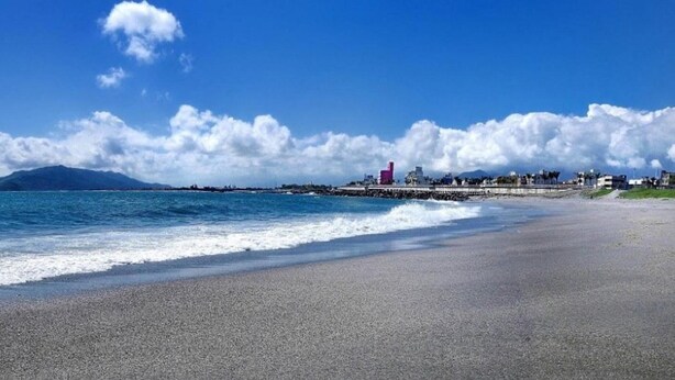 Ein Strand mit blauem Himmel, Wellen und einer entfernten Stadtlandschaft mit Gebäuden und einem Berg.