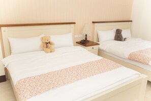 Ein Hotelzimmer mit zwei Betten, beide mit weißem Kopfteil und einem floralen Bettbezug. Auf einem Bett liegt ein Teddybär und auf dem Nachttisch steht eine Lampe.