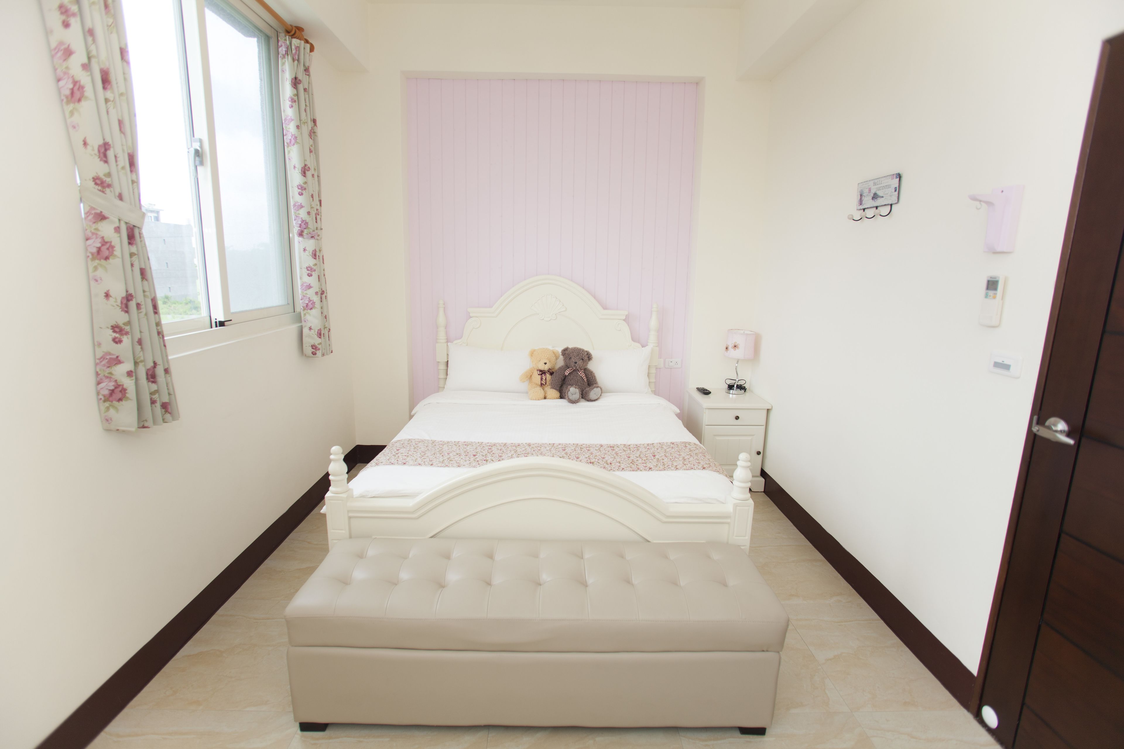 A bedroom with a white bed, a bedside table, a nightstand, and a window with floral curtains.