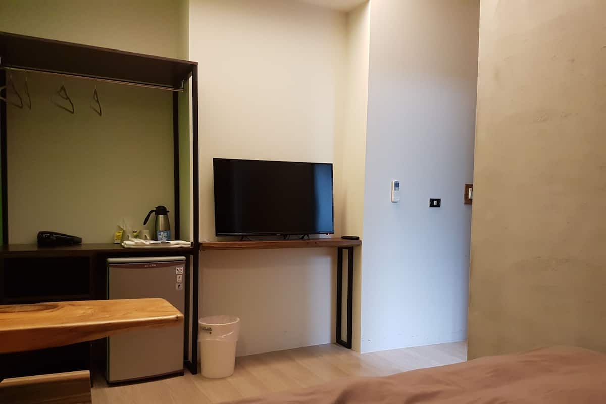 A compact hotel room with a bed, a small desk, a television, a mini-fridge, and a trash bin.