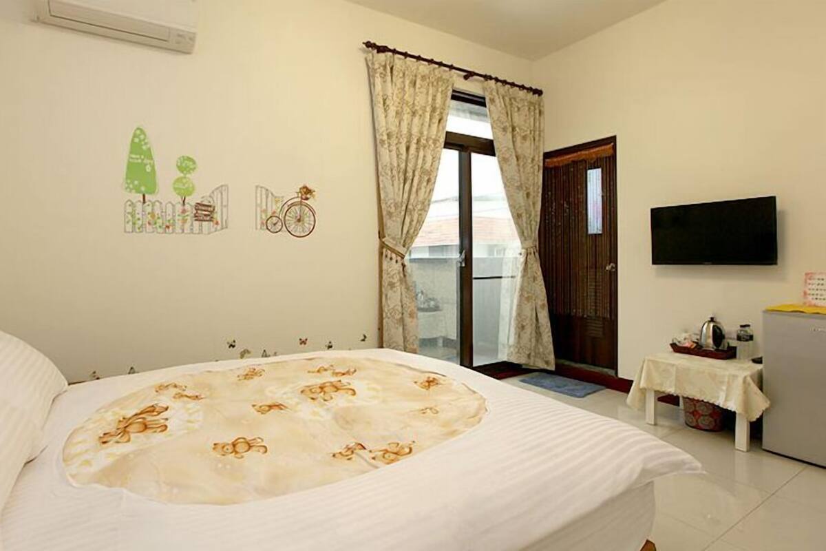Quarto de hotel com cama, televisão, mesinha com chaleira e janela com cortinas.