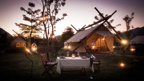 A rustic outdoor dining area with a table set for two, surrounded by wooden chairs, under a large tent with a view of the evening sky. - Lala Mukha Tented Resort Khao Yai (Pak Chong)