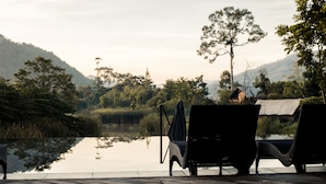 A serene lakeside setting with two wicker lounge chairs facing the water, surrounded by lush greenery and hills in the background. - Lala Mukha Tented Resort Khao Yai (Pak Chong)