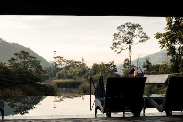 A serene lakeside setting with two wicker lounge chairs facing the water, surrounded by lush greenery and hills in the background. - Lala Mukha Tented Resort Khao Yai (Pak Chong)