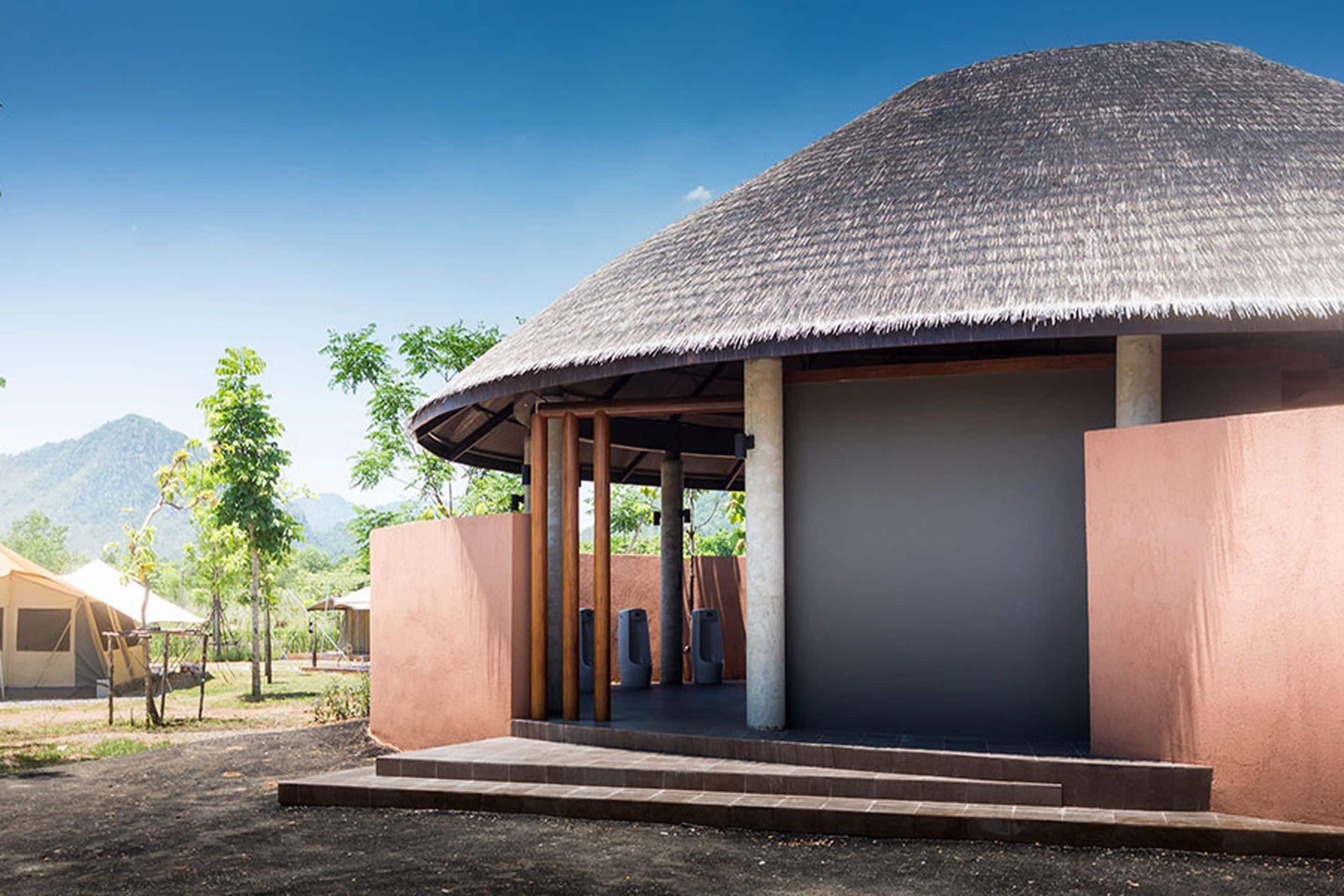 A thatched-roof structure with a covered porch and steps leading up to it, surrounded by a natural landscape and other buildings.