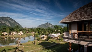 A resort with thatched-roof cottages by a lake, surrounded by mountains. - Lala Mukha Tented Resort Khao Yai (Pak Chong)