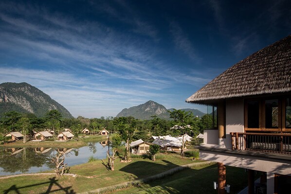 A resort with thatched-roof cottages by a lake, surrounded by mountains. - Lala Mukha Tented Resort Khao Yai (Pak Chong)