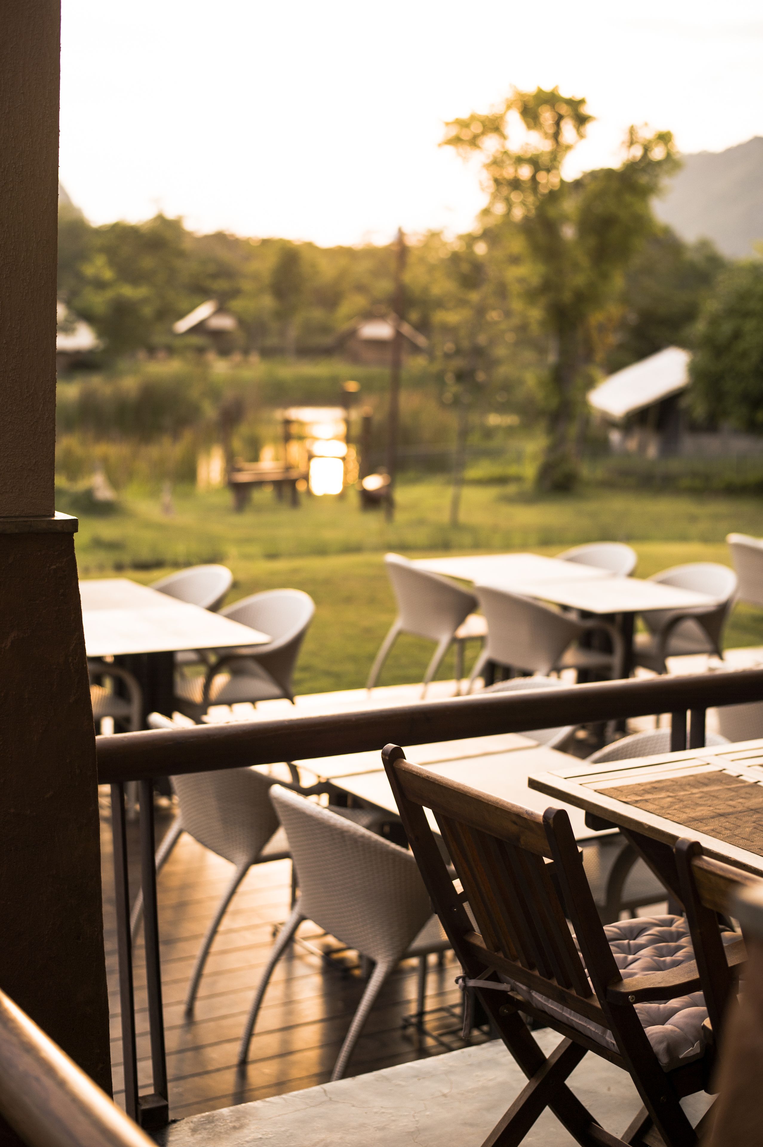An outdoor seating area with tables and chairs, a wooden deck, and a view of a grassy field and trees in the background.