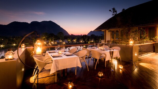 An outdoor dining area with tables set for a meal, surrounded by lanterns and overlooking a body of water with mountains in the background. - Lala Mukha Tented Resort Khao Yai (Pak Chong)
