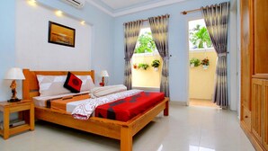 A hotel room with a wooden bed, two bedside tables, a nightstand, a window with curtains, and a view of greenery outside. - Lila Homestay Hoi An (Da Nang)