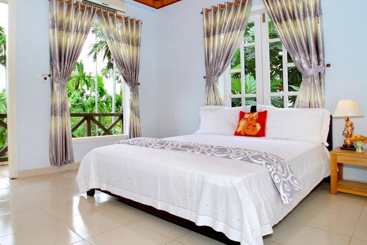 A bedroom with a large bed, curtains, a nightstand, and a view of greenery outside.
