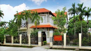A two-story building with a red-tiled roof, surrounded by a black fence and palm trees. - Lila Homestay Hoi An (Da Nang)
