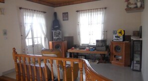 Ein Wohnzimmer mit einer Holztreppe, zwei mit Vorhängen versehenen Fenstern, einer Gitarre und einem Schreibtisch mit Audioausrüstung.