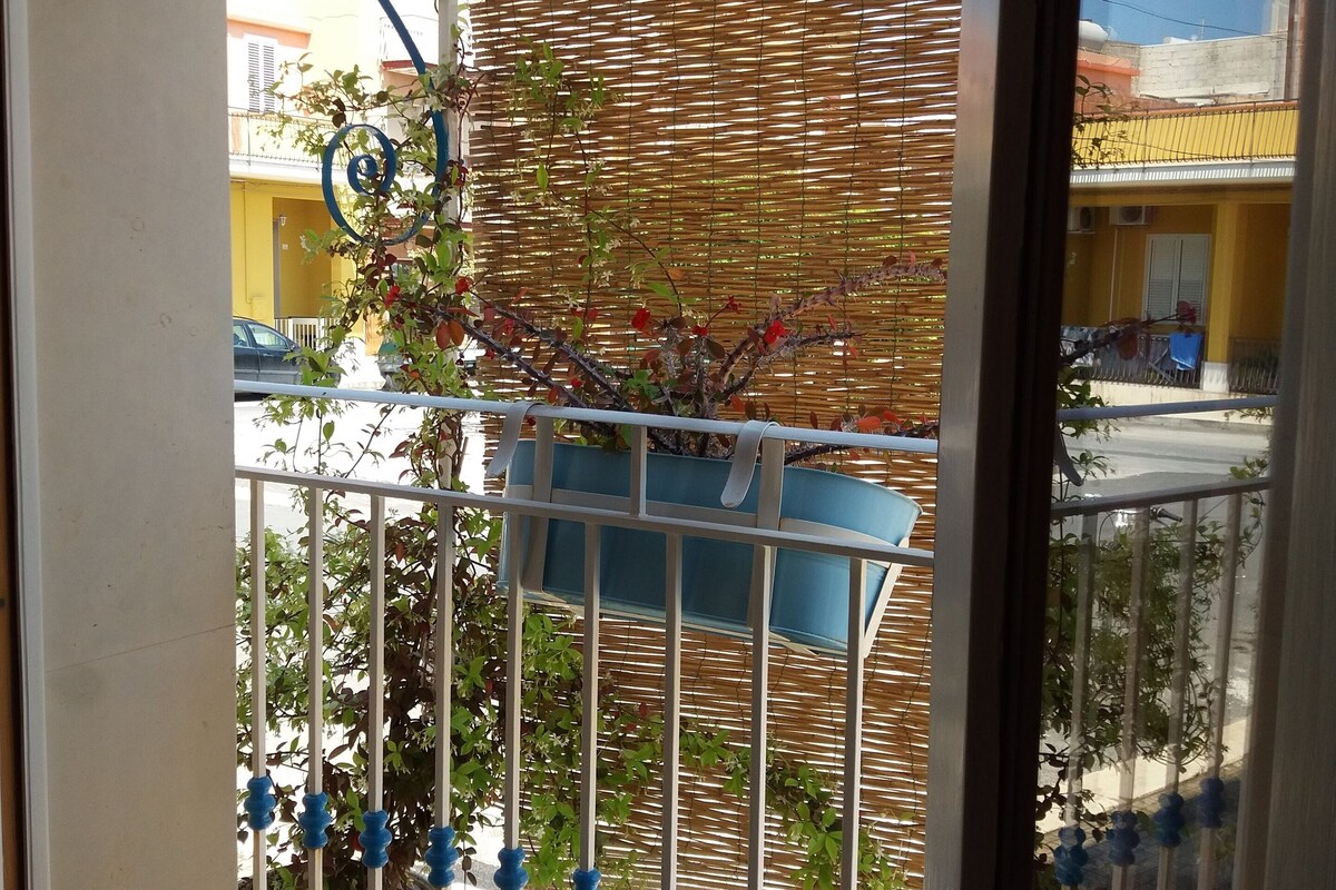 Un balcón con celosía de mimbre, baranda de metal con detalles azules y vista a una calle con edificios y coches aparcados.