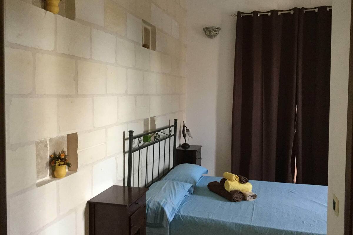 A bedroom with a bed, a nightstand, a wall-mounted shelf with a plant, a small window, and a curtain.