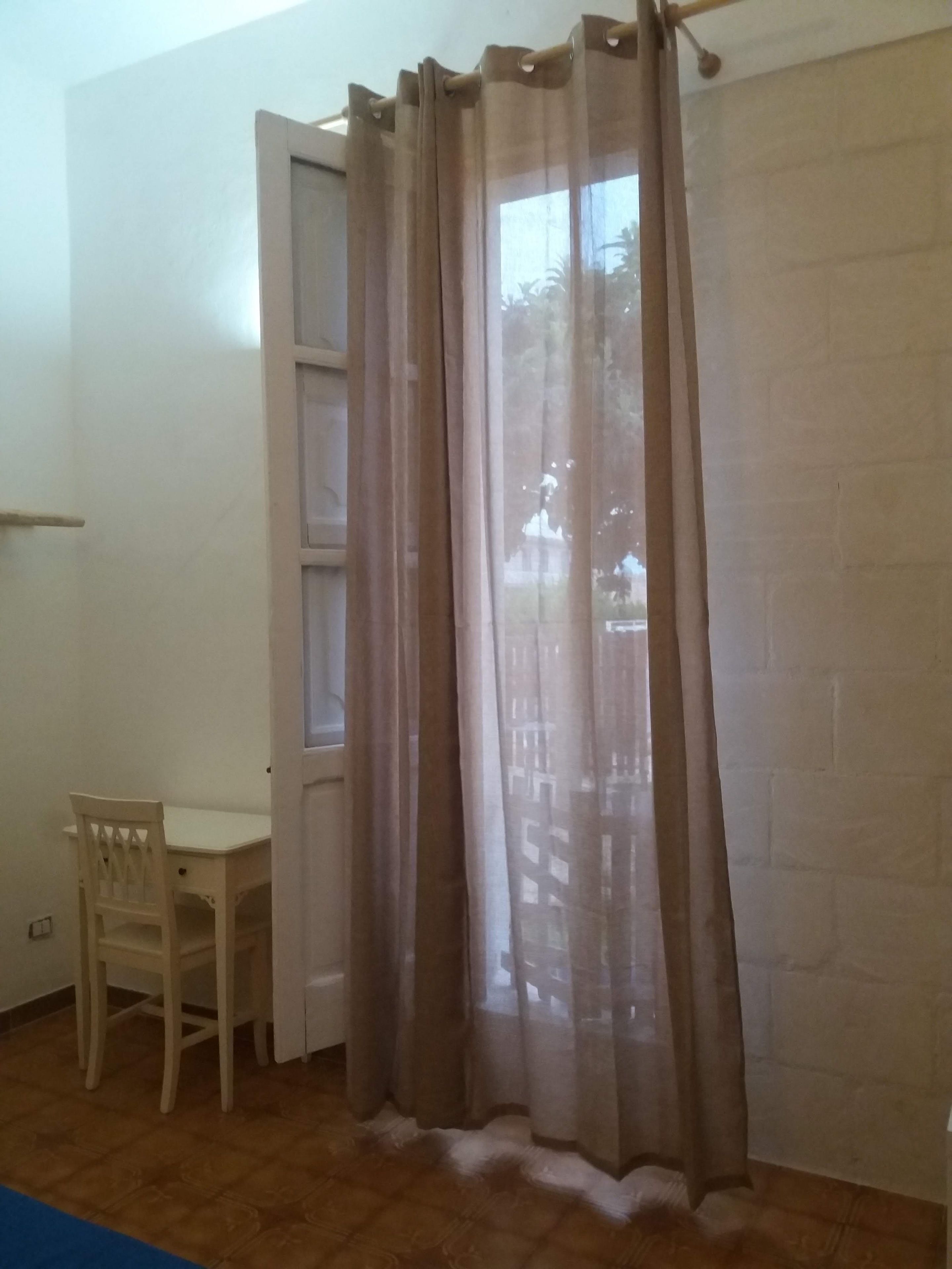 Quarto com escrivaninha branca, cadeira e janela com vista para móveis de exterior.
